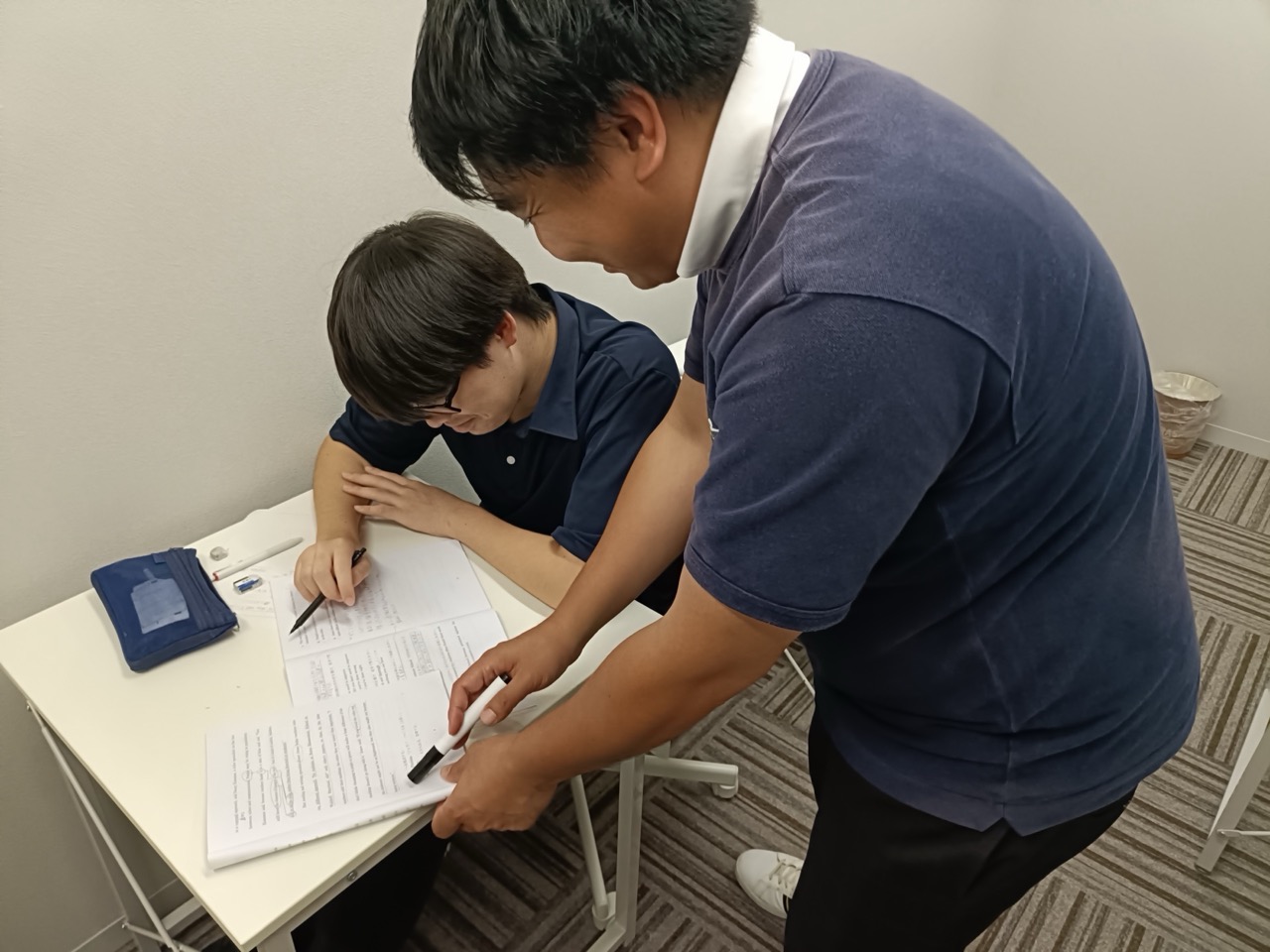 男性講師が生徒の机に立ち寄り、プリントや参考書を一緒に見ながら個別に指導する横浜予備校の授業風景