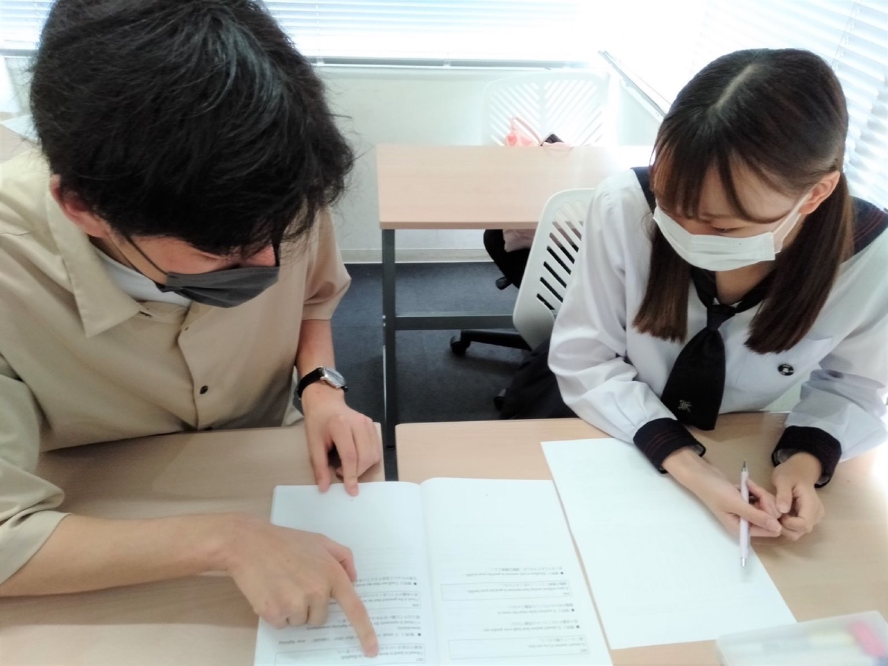 マスク姿のコーチがノートを示しながら、生徒と並んで学習計画を確認する様子
