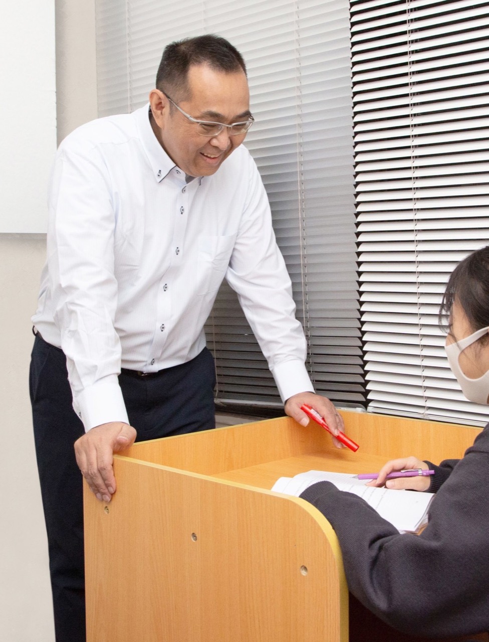 メガネをかけた白いシャツの男性講師が、机を挟んで生徒のノートを指しながら丁寧に解説する様子